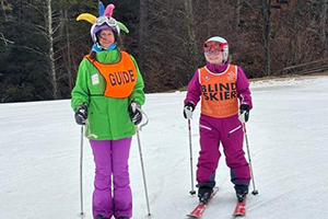 two skiiers wearing orange vests; one says 'Guide'. The other says 'blind skiier'