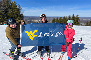 three skiiers on top of a mountain holding up a WVU flag that says Let's Go!